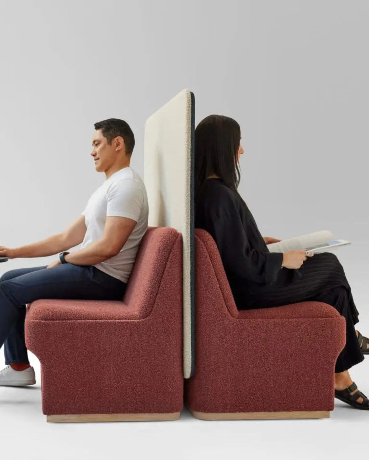 Two people sitting on red chairs with a partition between them, one using a laptop and the other reading.
