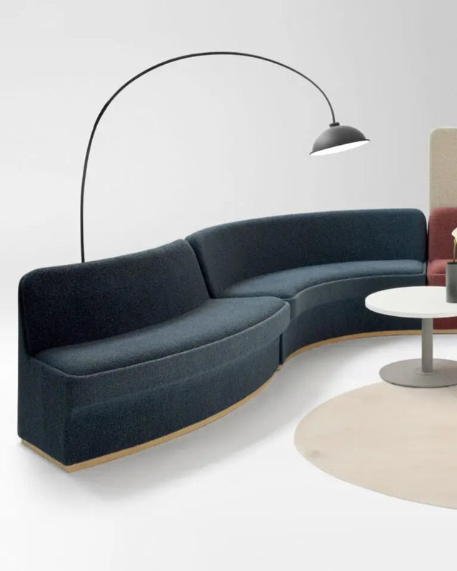 A modern modular lounge set featuring a curved dark navy blue sectional sofa, a terracotta red L-shaped sectional sofa, and a beige upholstered backrest panel, arranged around a round beige rug with a small white coffee table and a black floor lamp on a light gray background.