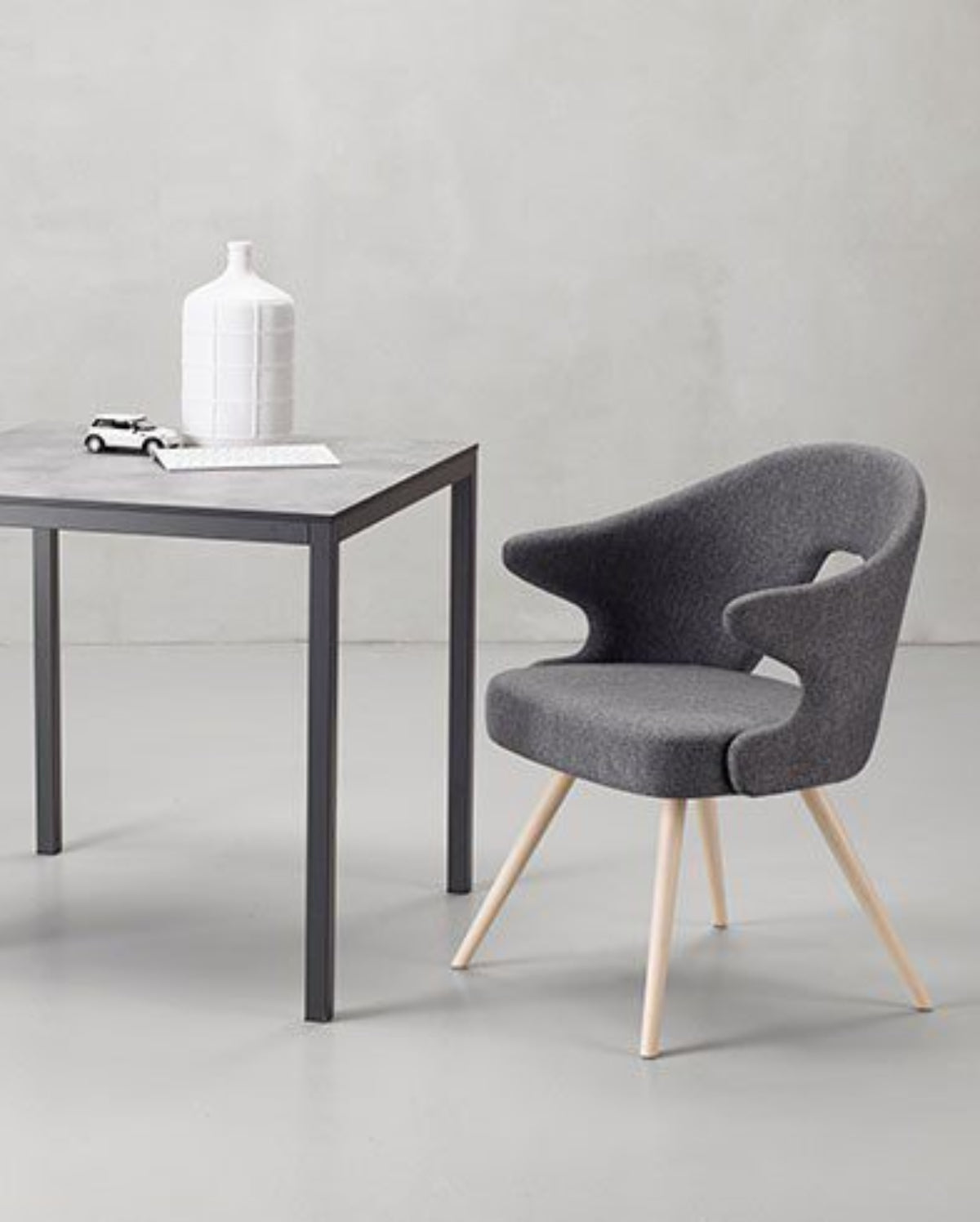 A modern dining set featuring a square gray table with dark metal legs and two matching gray upholstered chairs with light wood legs, arranged on a light gray floor against a textured gray wall.