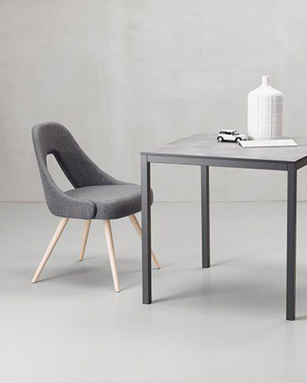 A modern dining set featuring a square gray table with dark metal legs and two matching gray upholstered chairs with light wood legs, arranged on a light gray floor against a textured gray wall.