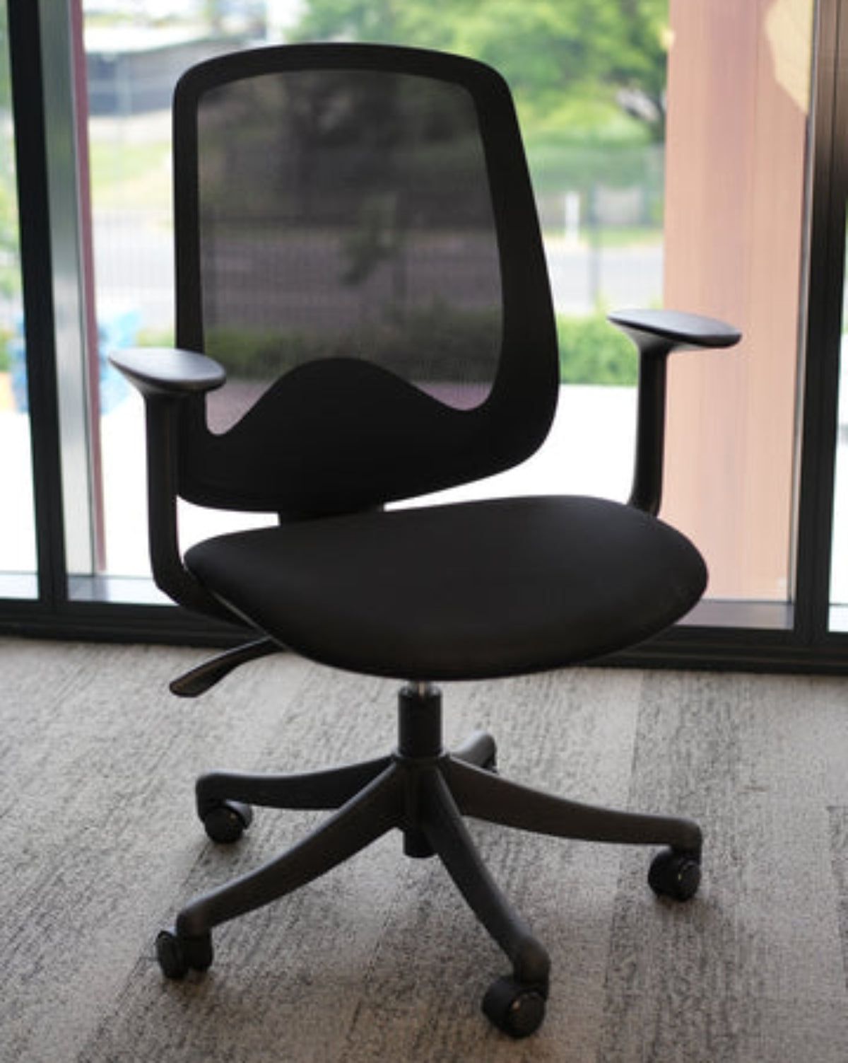 A black mesh office chair with adjustable armrests and a five-wheel base, positioned in front of a large window showing a blurred outdoor background.