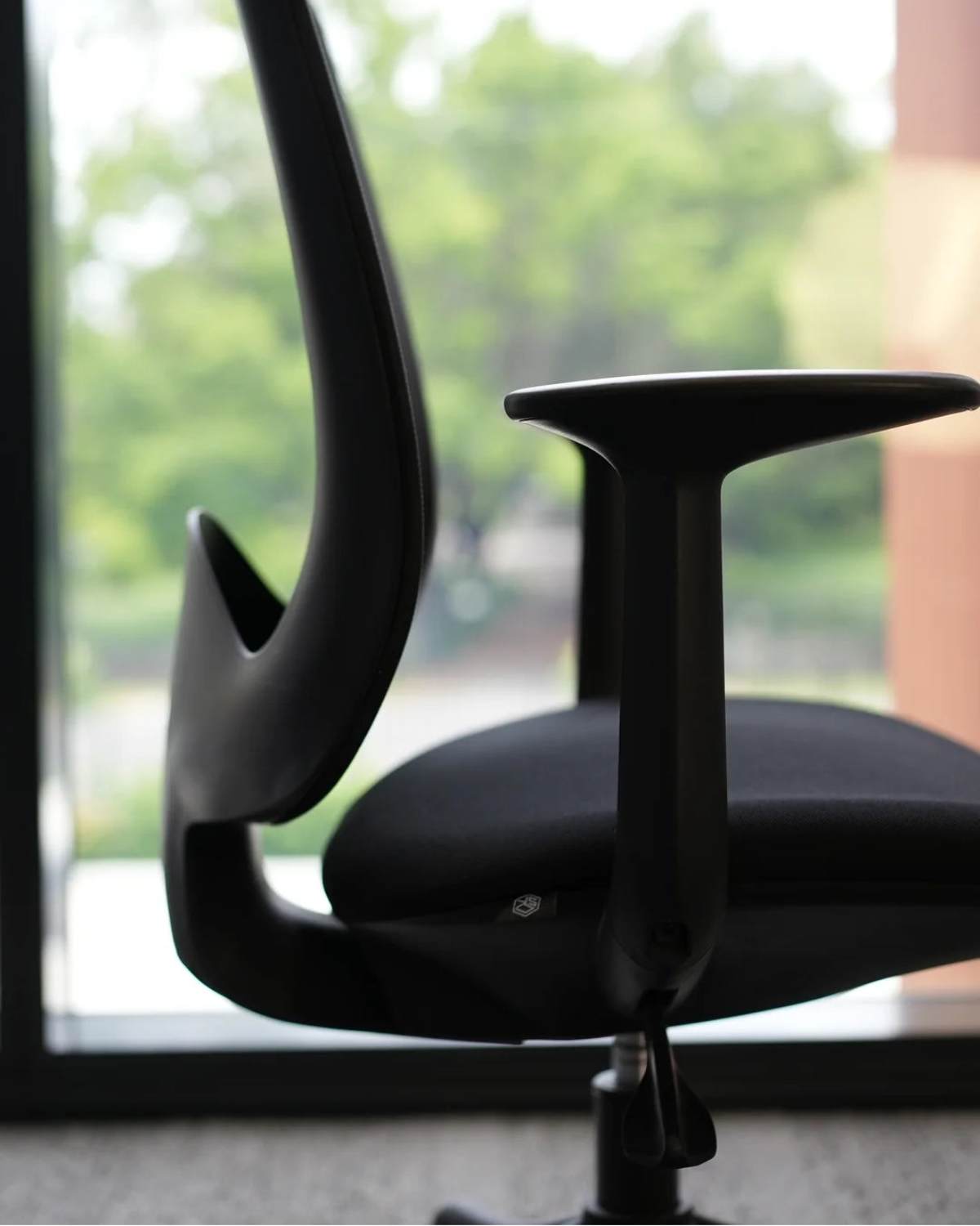 A modern black ergonomic office chair with a curved high-back support and padded seat, shown in profile near a window with a blurred outdoor green background.