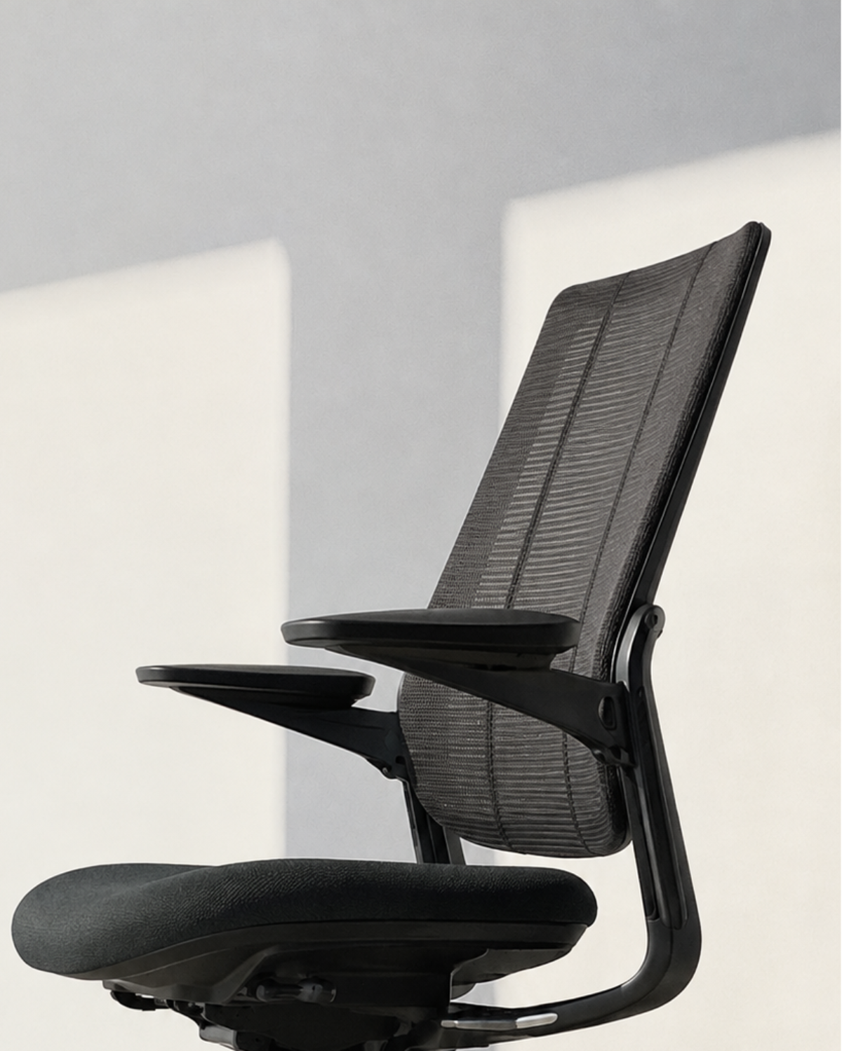A modern black mesh office chair with adjustable armrests, a padded seat, and a five-wheel base, positioned against a minimalist white wall with soft shadows.