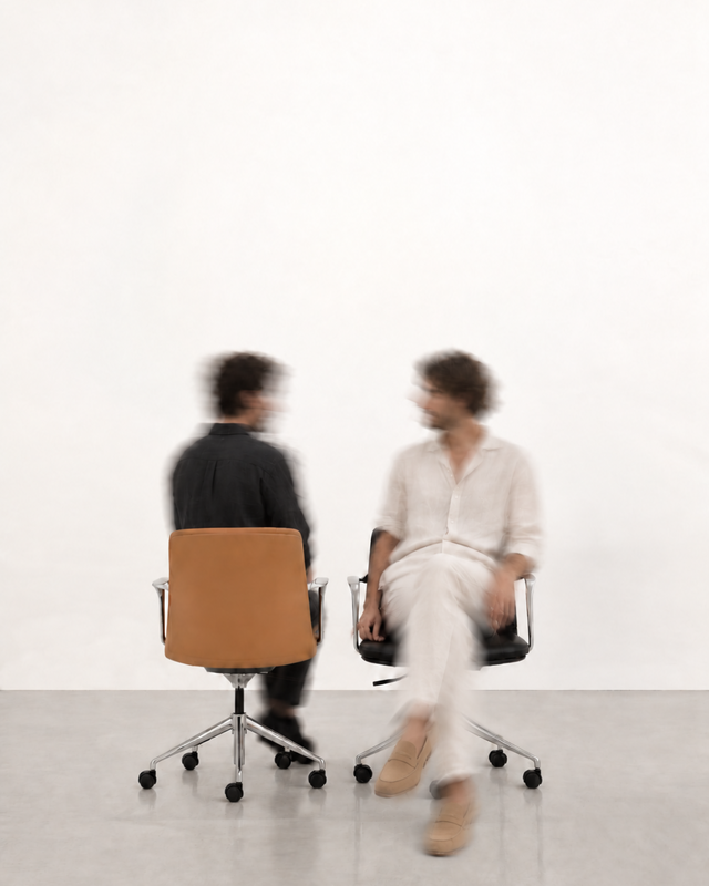 Two blurred people seated in office chairs in a minimalist white room with a gray floor.