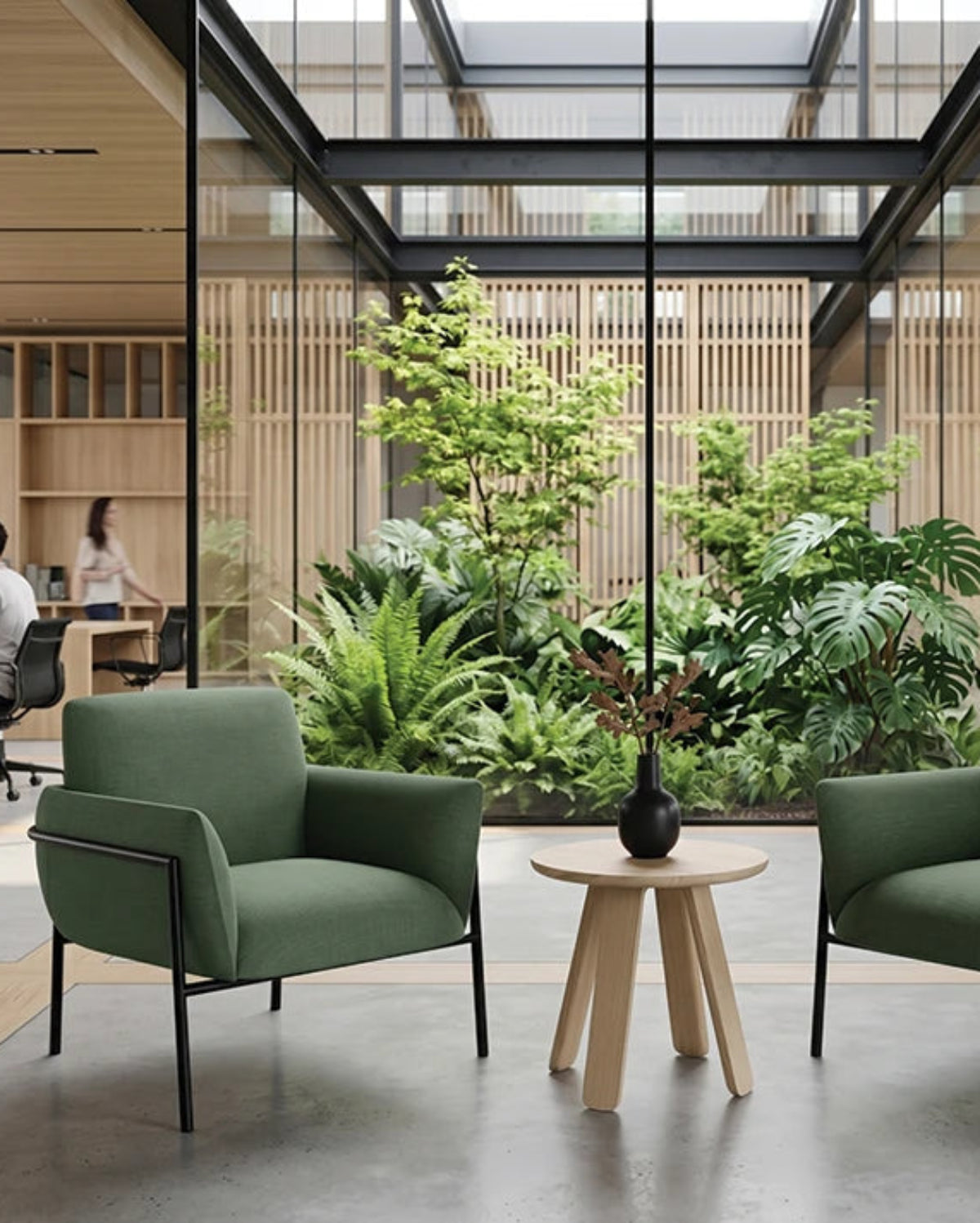 Two green upholstered armchairs with black metal frames flank a small round wooden side table in a modern office lobby. Large glass walls and a central indoor garden are visible in the background, with office workers seated at desks.