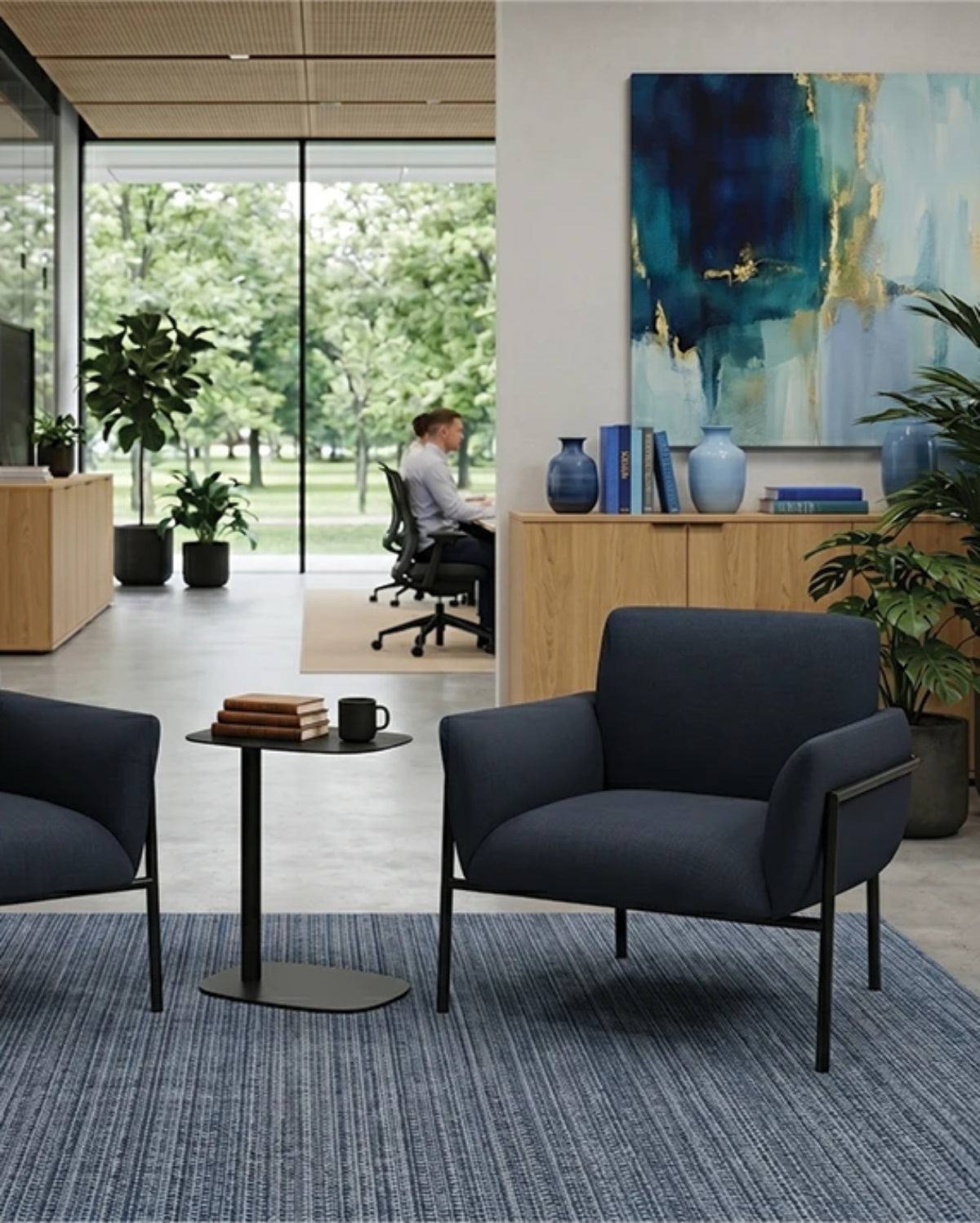 Two dark blue upholstered office chairs with black metal frames arranged around a small round side table in a modern office lobby. A blue patterned area rug covers the floor, and a woman sits at a desk in the background near large windows and an abstract painting.
