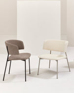 Two modern accent chairs in a minimalist studio setting. The chair on the left is upholstered in a textured taupe fabric with black metal legs.