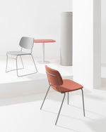 A modern minimalist interior scene with two chairs and a small round table on a white platform. The foreground chair has a terracotta orange upholstered seat and back with thin gray metal legs.