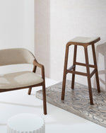 A modern wooden bar stool with a light beige upholstered seat and back, shown in a minimalist interior setting next to a matching armchair.