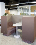 A modern office seating area featuring brown acoustic booth partitions with upholstered bench seating and a small white round table in the center.