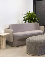 A modern two-seat sofa upholstered in light gray textured fabric with beige side panels, shown in a minimalist office or lounge setting with a matching ottoman, side table, and green plant.