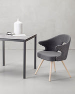A modern dining set featuring a square gray table with dark metal legs and two matching gray upholstered chairs with light wood legs, arranged on a light gray floor against a textured gray wall.
