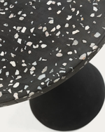 Close-up of a round black table with a white terrazzo speckled pattern on the tabletop and a black circular base, photographed on a white background.