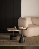 Two modern round side tables styled in a minimalist living room next to a cream bouclé armchair. One table has a light wood top with dark speckles and a black pedestal base, while the other has a dark stone top and a black conical base.