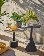 Two round tables with plants and a glass on a concrete floor.