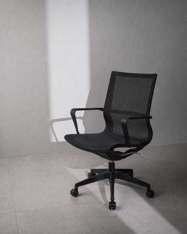 A black mesh office chair with armrests and a five-wheel base, positioned against a minimalist gray wall and tiled floor with dramatic diagonal shadows.