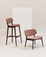 Three upholstered dining or bar stools in a minimalist studio setting. The stools feature textured dusty rose pink fabric seats and backs, with dark brown and black wooden legs on the left and center, and light beige wood legs on the right.