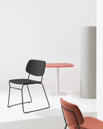 A modern interior scene with two chairs and a small table on white platform steps. The foreground chair has a rust-orange upholstered seat and back with thin black metal legs.