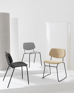 Three modern accent chairs with black metal wire frames displayed on white platform steps against a minimalist white studio background. The chairs feature different upholstery colors: one black, one gray, and one beige.