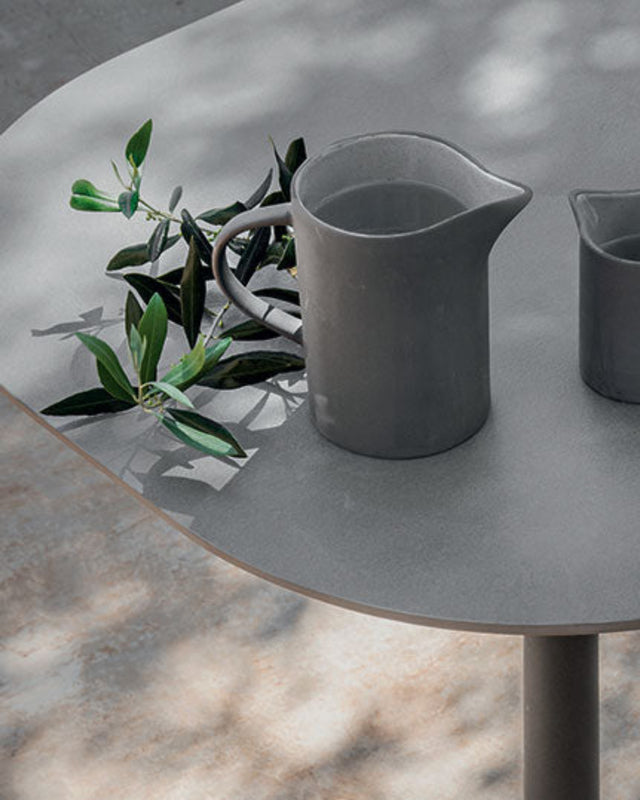 A modern round outdoor table with a gray stone-look top and base, paired with woven light wood chairs. On the table are two matte gray ceramic pitchers and a small branch with green leaves, set on a concrete patio with dappled sunlight and shadows.