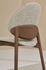 A close-up of a modern dining chair featuring a textured cream and brown woven fabric upholstery and a curved medium-brown wooden frame, set against a minimalist beige background.
