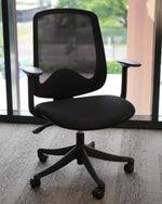 A black mesh office chair with adjustable armrests and a five-wheel base, positioned in front of a large window showing a blurred outdoor background.