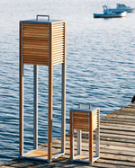 Ginger tall and table lamps placed outdoors on a wooden deck next to some water, with some boats in the background.