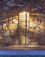 Ginger Tall Lamp placed outside, the time is dusk and the lamp is lighting up the stone wall behind it.