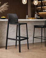 A modern black mesh high bar stool with a square footrest, positioned in front of a white marble kitchen island in a contemporary kitchen setting.
