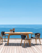 Outdoor dining set with wooden table and chairs on a deck overlooking the ocean.
