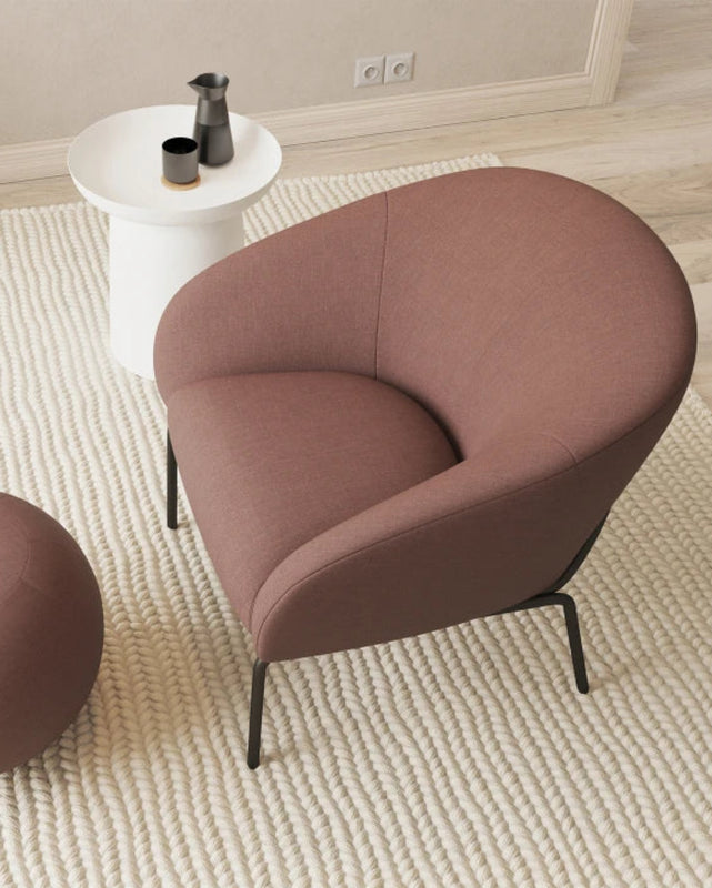 A modern upholstered armchair in a dusty rose-brown fabric with black metal legs, shown alongside a matching round ottoman on a textured cream rug in a styled interior setting.