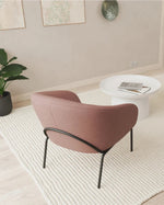 A modern living room scene featuring a dusty rose upholstered armchair with black metal legs, a white round coffee table, two spherical poufs (one dusty rose, one light gray), and a large cream-colored textured area rug on a light wood floor.