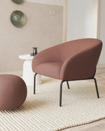 A modern upholstered armchair in a dusty rose-brown fabric with black metal legs, shown alongside a matching round ottoman in a styled living room setting.