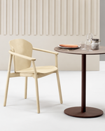 A modern three-piece dining set featuring two light beige armchairs and a round dark brown pedestal table, styled with a plate, glass, and metal can on a neutral studio background.