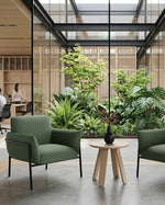 Two green upholstered armchairs with black metal frames flank a small round wooden side table in a modern office lobby. Large glass walls and a central indoor garden are visible in the background, with office workers seated at desks.