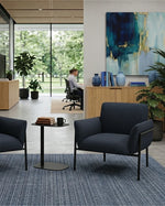 Two dark blue upholstered office chairs with black metal frames arranged around a small round side table in a modern office lobby. A blue patterned area rug covers the floor, and a woman sits at a desk in the background near large windows and an abstract painting.