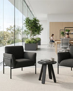 Two dark gray lounge armchairs with black metal frames and a small round black side table, arranged in a bright modern office lobby with large windows, plants, and seated workers in the background.