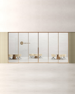 A modern glass office partition wall with slim gold-toned metal frames, shown in a minimalist interior setting. The glass panels reflect meeting tables and chairs, and the wall features vertical striped side panels in beige and olive tones.