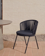 Black wicker chair with cushion on a tiled floor against a brick wall.