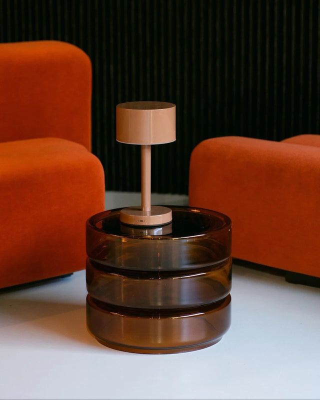 A modern side table lamp with a translucent amber-brown glass cylindrical base and a minimalist beige metal lamp post and shade, set between two orange armchairs against a dark textured wall.