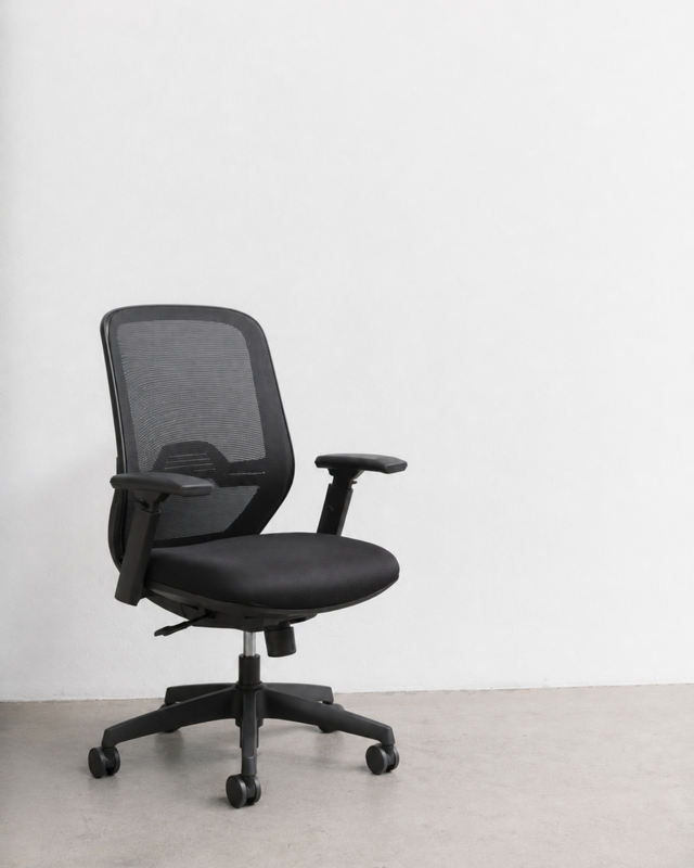 A modern black mesh office chair with adjustable armrests and a five-star wheeled base, positioned against a plain white wall and light gray concrete floor.