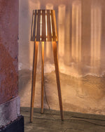 Woody Floor Lamp inside lit up, on a wooden deck floor with the light from the lamp reflecting on the terracotta walls.