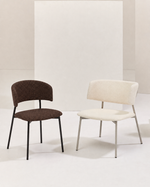 Two modern accent chairs with curved upholstered backs and seats on slim metal legs, shown on a clean studio set. One chair is dark brown and the other is off-white/cream, both against a minimalist beige backdrop.