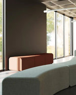 A modern curved upholstered seating bench in a light sage green fabric, shown in a contemporary interior space with large windows, a dark wall, and another rust-colored bench in the background.