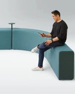A man sits on a curved modular upholstered seating unit in a bright studio setting, holding a phone. The seating is a muted teal-blue color, with a small round black side table attached near one corner.