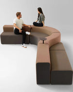 A curved modular upholstered seating unit in warm taupe and dark charcoal brown, shown with two people sitting on it in a clean studio setting.