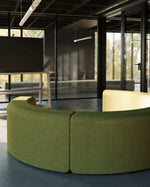A curved modular upholstered sofa in olive green fabric, shown in a modern office interior with large glass walls and dark ceiling panels.