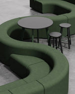 A modern curved upholstered seating nook in dark green fabric, featuring a low round table and two bar stools on a light gray concrete floor.